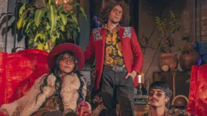 Three people dressed in eclectic, retro-style clothing pose in a vintage room with red furniture and plants.