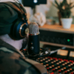 A person wearing headphones and a beanie speaks into a microphone in a recording studio, surrounded by audio equipment and a potted plant, producing content for an artificial streaming platform.