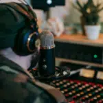 A person wearing headphones and a beanie speaks into a microphone in a recording studio, surrounded by audio equipment and a potted plant, producing content for an artificial streaming platform.