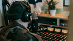 A person wearing headphones and a beanie speaks into a microphone in a recording studio, surrounded by audio equipment and a potted plant, producing content for an artificial streaming platform.