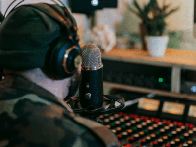 A person wearing headphones and a beanie speaks into a microphone in a recording studio, surrounded by audio equipment and a potted plant, producing content for an artificial streaming platform.