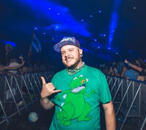 A person in a green dinosaur shirt and cap poses confidently with a hand gesture, embodying client marketing energy amidst the vibrant concert crowd and dazzling stage lights.