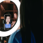 A woman records herself using a smartphone mounted on a tripod with a ring light, capturing content for her latest TikTok. She is in a room with shelves in the background.