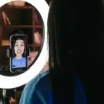 A woman records herself using a smartphone mounted on a tripod with a ring light, capturing content for her latest TikTok. She is in a room with shelves in the background.