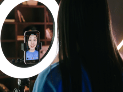 A woman records herself using a smartphone mounted on a tripod with a ring light, capturing content for her latest TikTok. She is in a room with shelves in the background.