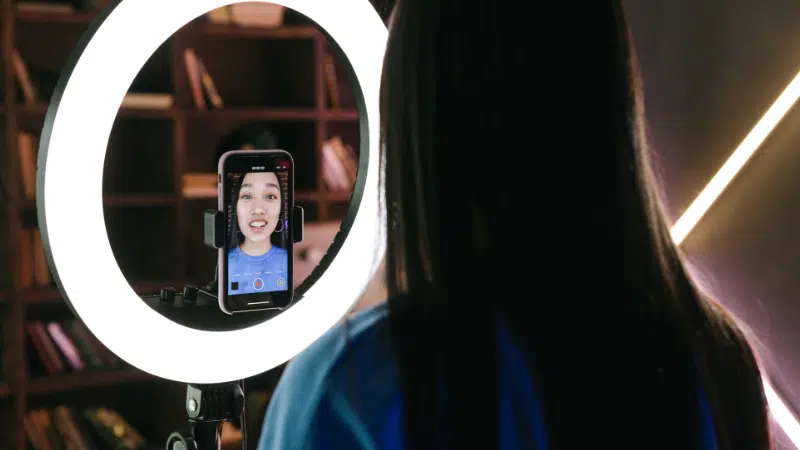 A woman records herself using a smartphone mounted on a tripod with a ring light, capturing content for her latest TikTok. She is in a room with shelves in the background.