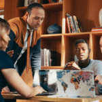 Four people in a meeting room, including new interns, gathered around a table with a laptop, discussing and working together. Books and decorative items adorn the shelves in the background, setting an inspiring atmosphere.