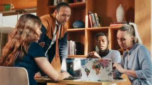 Four people in a meeting room, including new interns, gathered around a table with a laptop, discussing and working together. Books and decorative items adorn the shelves in the background, setting an inspiring atmosphere.