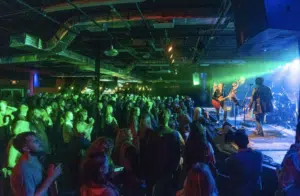 A band performs on stage in a dimly lit venue. An audience stands watching, captivated by the live music. Green and blue lights illuminate the scene, creating a vibrant atmosphere.