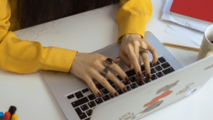 A person in a yellow shirt types on a sticker-adorned laptop, surrounded by documents, a mug, and writing tools on a white table. Perhaps they're crafting an email in Spanish or jotting down notes for their next big project.