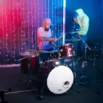 A drummer plays a drum kit on stage with colorful lights behind. Nearby, a person in a hoodie adjusts a camera on a tripod, capturing the moment for YouTube.