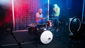 A drummer plays a drum kit on stage with colorful lights behind. Nearby, a person in a hoodie adjusts a camera on a tripod, capturing the moment for YouTube.