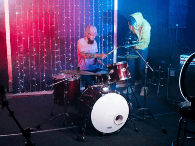 A drummer plays a drum kit on stage with colorful lights behind. Nearby, a person in a hoodie adjusts a camera on a tripod, capturing the moment for YouTube.