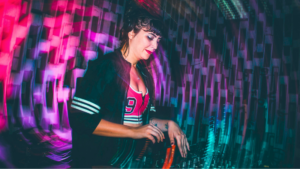 A woman in a sports-style jacket uses a DJ mixer, with colorful, blurred lights and patterned walls in the background, embodying the energy of women led music.