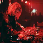 DJ in a dark, red-lit club mixes tracks on sound equipment, wearing large headphones.