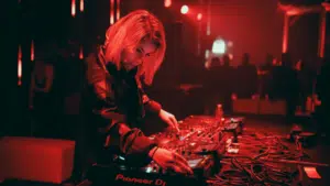 DJ in a dark, red-lit club mixes tracks on sound equipment, wearing large headphones.