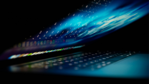 A tech-inspired laptop partially closed, its screen glowing in the dark and casting colorful icons and reflections across the keyboard.