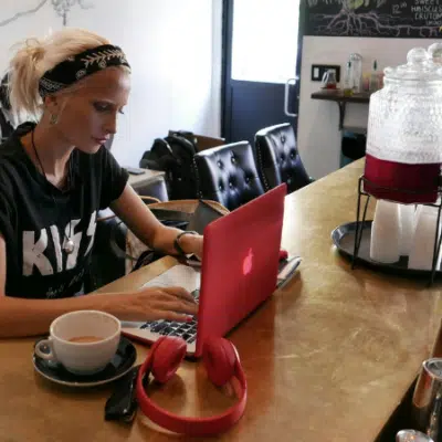 A person with blonde hair and a headband works on a red laptop at a café counter, with a coffee cup and red headphones nearby.