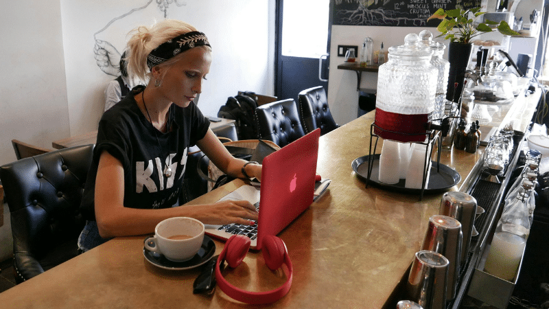 A person with blonde hair and a headband works on a red laptop at a café counter, with a coffee cup and red headphones nearby.