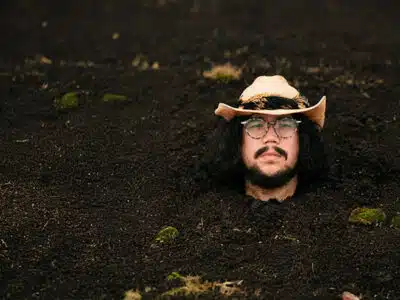 A man wearing glasses and a cowboy hat is buried up to his neck in dark soil, with only his head visible above the ground.