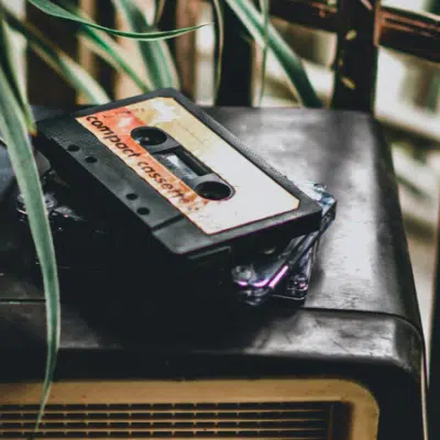 A cassette tape labeled "compact cassette" rests on top of a vintage radio, with plant leaves partially visible in the foreground.