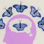 A purple silhouette of a human head with a butterfly inside its brain area, symbolizing mental health, is surrounded by five blue butterflies arranged in an arc above it.
