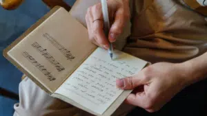 A person writes lyrics in a notebook with a pen; the notebook page on the left shows handwritten music notes on a staff.