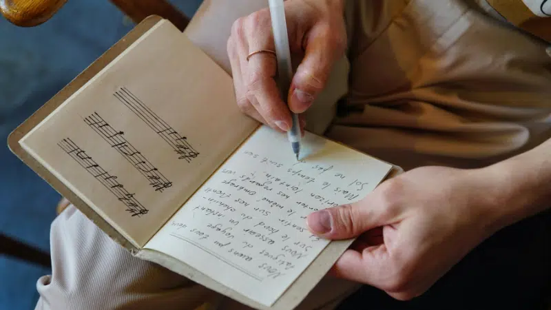 A person writes lyrics in a notebook with a pen; the notebook page on the left shows handwritten music notes on a staff.