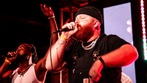 Two men perform on stage with microphones, one wearing a black beanie and overalls, the other in a white tank top, against a backdrop of colorful lights. Their dynamic presence shines as they entertain the crowd, hoping their passion for music will secure future funding for bigger shows.