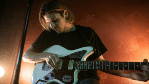Publishing 101 for Indie Labels: What You Need to Know to Do It Right A person with long hair plays an electric guitar in a dimly lit room with reddish-brown walls, perhaps lost in creative thought before publishing their next song.