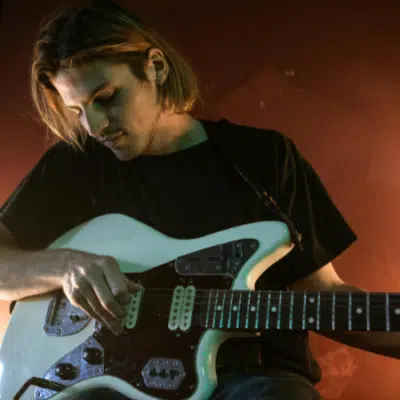A person with long hair plays an electric guitar in a dimly lit room with reddish-brown walls, perhaps lost in creative thought before publishing their next song.
