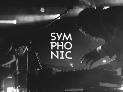 Amidst the dazzling lights, a musician stands silhouetted against an electrifying backdrop labeled "SYMPHONIC," captivating the audience with each note. This mesmerizing performance doubles as a grand promotion of their latest transformative soundscapes.