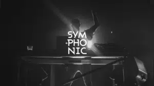 A DJ stands behind a table with tech equipment on stage under dramatic lighting, with the word "SYMPHONIC" displayed prominently in bold white letters.