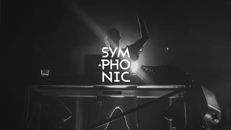A DJ stands behind a table with tech equipment on stage under dramatic lighting, with the word "SYMPHONIC" displayed prominently in bold white letters.