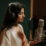 A woman with long dark hair wearing headphones stands in front of a microphone in a recording studio, ready to deliver a masterclass in vocal performance.