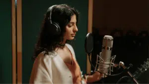 A woman with long dark hair wearing headphones stands in front of a microphone in a recording studio, ready to deliver a masterclass in vocal performance.