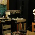 Room with vintage audio equipment, including reel-to-reel tape recorders, turntables, and amplifiers—a haven for physical music enthusiasts—featuring shelves of vinyl records and a large painting of sailboats on the wall.