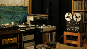Room with vintage audio equipment, including reel-to-reel tape recorders, turntables, and amplifiers—a haven for physical music enthusiasts—featuring shelves of vinyl records and a large painting of sailboats on the wall.