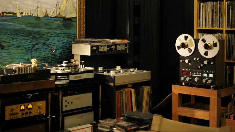 Room with vintage audio equipment, including reel-to-reel tape recorders, turntables, and amplifiers—a haven for physical music enthusiasts—featuring shelves of vinyl records and a large painting of sailboats on the wall.