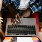 Three people sit together testing something on a laptop, with their hands visible on the keyboard and trackpad, viewed from above.