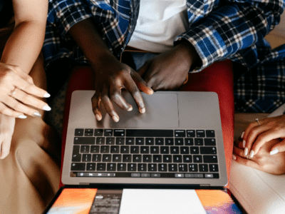 Three people sit together testing something on a laptop, with their hands visible on the keyboard and trackpad, viewed from above.