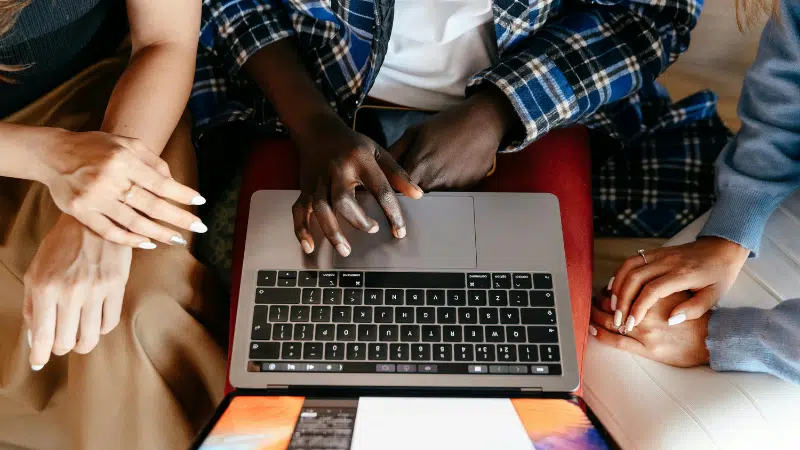 Three people sit together testing something on a laptop, with their hands visible on the keyboard and trackpad, viewed from above.