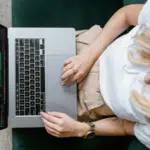 Person wearing an orange beanie and glasses sits on a green couch using a laptop, editing video footage for an online masterclass.