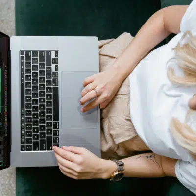 Person wearing an orange beanie and glasses sits on a green couch using a laptop, editing video footage for an online masterclass.