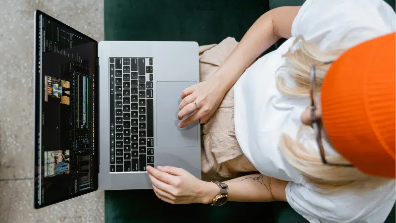 Person wearing an orange beanie and glasses sits on a green couch using a laptop, editing video footage for an online masterclass.