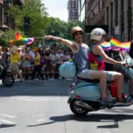 Two people ride a scooter and wave Pride rainbow flags in a city street parade, with a large crowd of spectators and other scooter riders in the background.