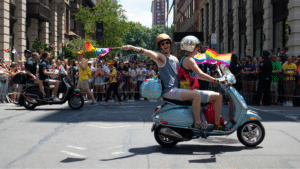 Two people ride a scooter and wave Pride rainbow flags in a city street parade, with a large crowd of spectators and other scooter riders in the background.