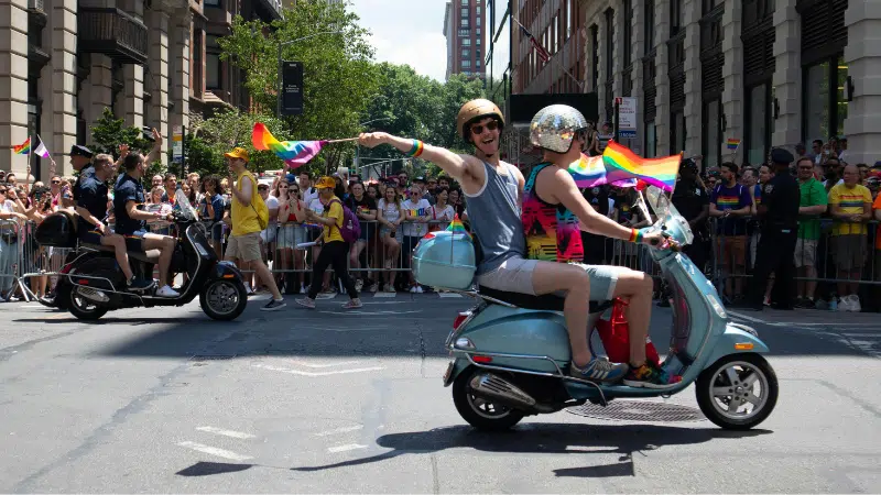 Two people ride a scooter and wave Pride rainbow flags in a city street parade, with a large crowd of spectators and other scooter riders in the background.