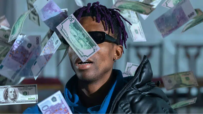 A person wearing sunglasses and a dark jacket is surrounded by various denominations of paper currency falling through the air, illustrating the chaos that can come from neglecting budgeting.