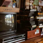 A vintage television and VCR sit in sync on a wooden cabinet next to a stack of VHS tapes and assorted books and papers.